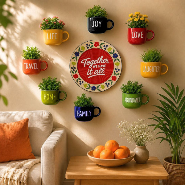 A styled interior wall with colorful mug-shaped planters labeled with words like JOY, LOVE, TRAVEL, HOME, FAMILY, and ABUNDANCE, plus a central decorative plate reading 'Together WE HAVE it all.' The scene includes a sofa, table, fruit, and potted plants in a warm, sunlit room.