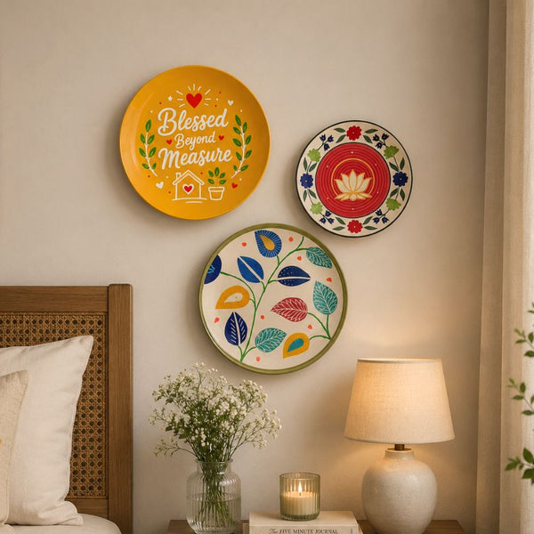 Three decorative wall plates with colorful floral and inspirational designs are displayed on a beige wall in a cozy bedroom setting next to a bed and a bedside table with a lamp and flowers.