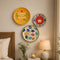 Three decorative wall plates with colorful floral and inspirational designs are displayed on a beige wall in a cozy bedroom setting next to a bed and a bedside table with a lamp and flowers.