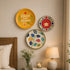 Three decorative wall plates with colorful floral and inspirational designs are displayed on a beige wall in a cozy bedroom setting next to a bed and a bedside table with a lamp and flowers.