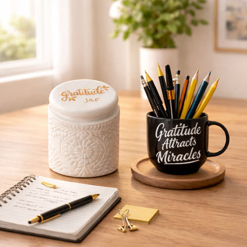 Desk setup with a gratitude jar, mug, notebook, and stationery on a wooden surface.