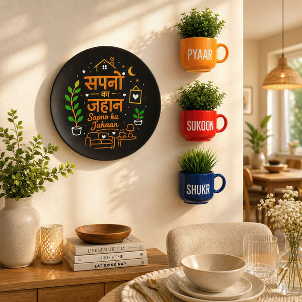 A round black decorative wall plaque with Hindi text and small illustrated icons hangs on a beige wall in a styled dining room. Three colorful mug-shaped plant holders labeled PYAAR, SUKOON, and SHUKUR are mounted to the right, along with potted green plants.