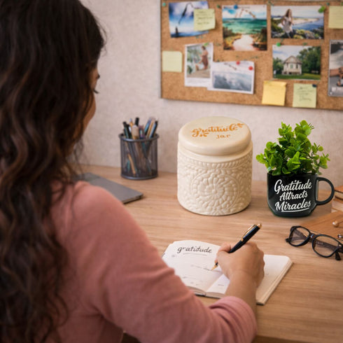 Person writing in a journal at a desk with a 'Gratitude' mug and decorative items.