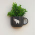 Black mug with white elephant design containing a green plant on a light background