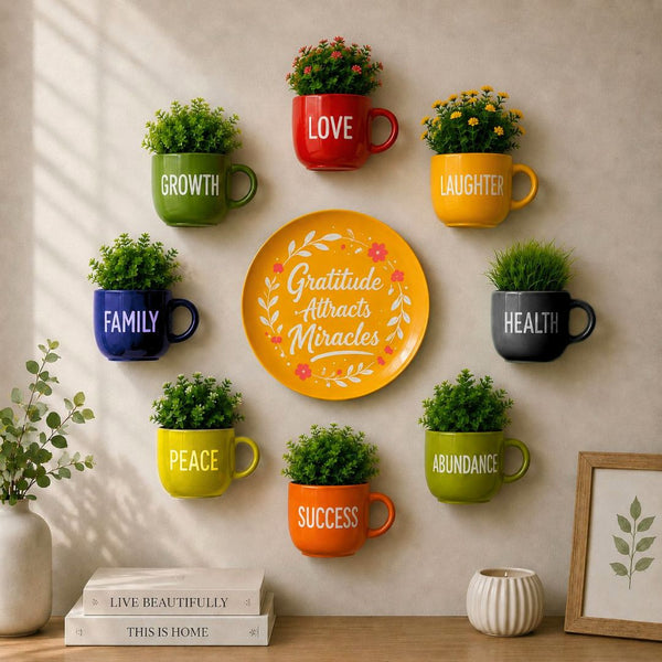 A styled wall arrangement of colorful ceramic mugs with plants, surrounding a central yellow plate that reads “Gratitude Attracts Miracles.” The mugs are labeled with words such as LOVE, LAUGHTER, FAMILY, HEALTH, PEACE, ABUNDANCE, SUCCESS, and GROWTH, displayed on a beige wall above a tabletop with books and decor.