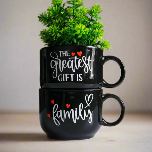 Two black mugs with white text and red hearts, one containing a small plant, on a neutral background.