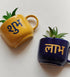 Two ceramic wall planters with one in yellow and the other in blue, each with a small plant inside and Hindi script on the side.